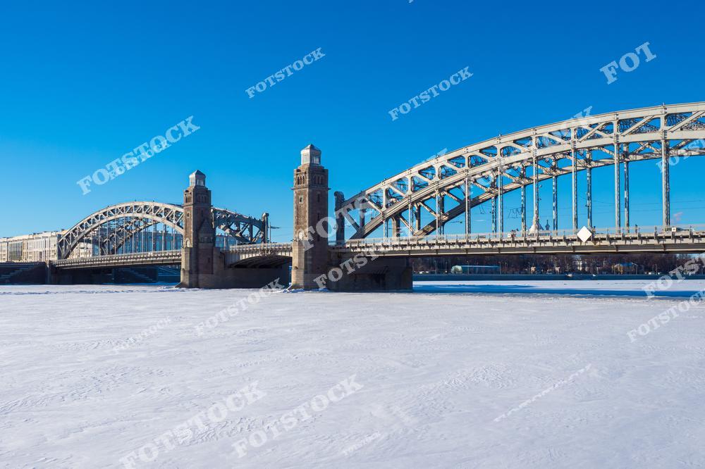 На фотографии изображен Большеохтинский мост, один из самых известных мостов Санкт-Петербурга. Массивные башни и металлические арки моста возвышаются над заснеженной поверхностью замерзшей Невы. Яркое солнце и безоблачное небо придают пейзажу свежесть и чистоту. Мост является не только важным инженерным сооружением, но и символом архитектурного мастерства.